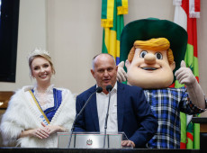 
Alceu Gilmar Moretti discursa na tribuna, acompanhado de Anna Letícia Hornburg e do mascote Hans.