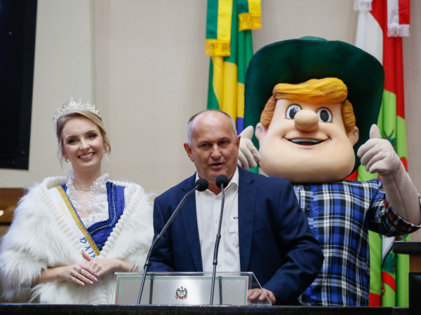 
Alceu Gilmar Moretti discursa na tribuna, acompanhado de Anna Letícia Hornburg e do mascote Hans.