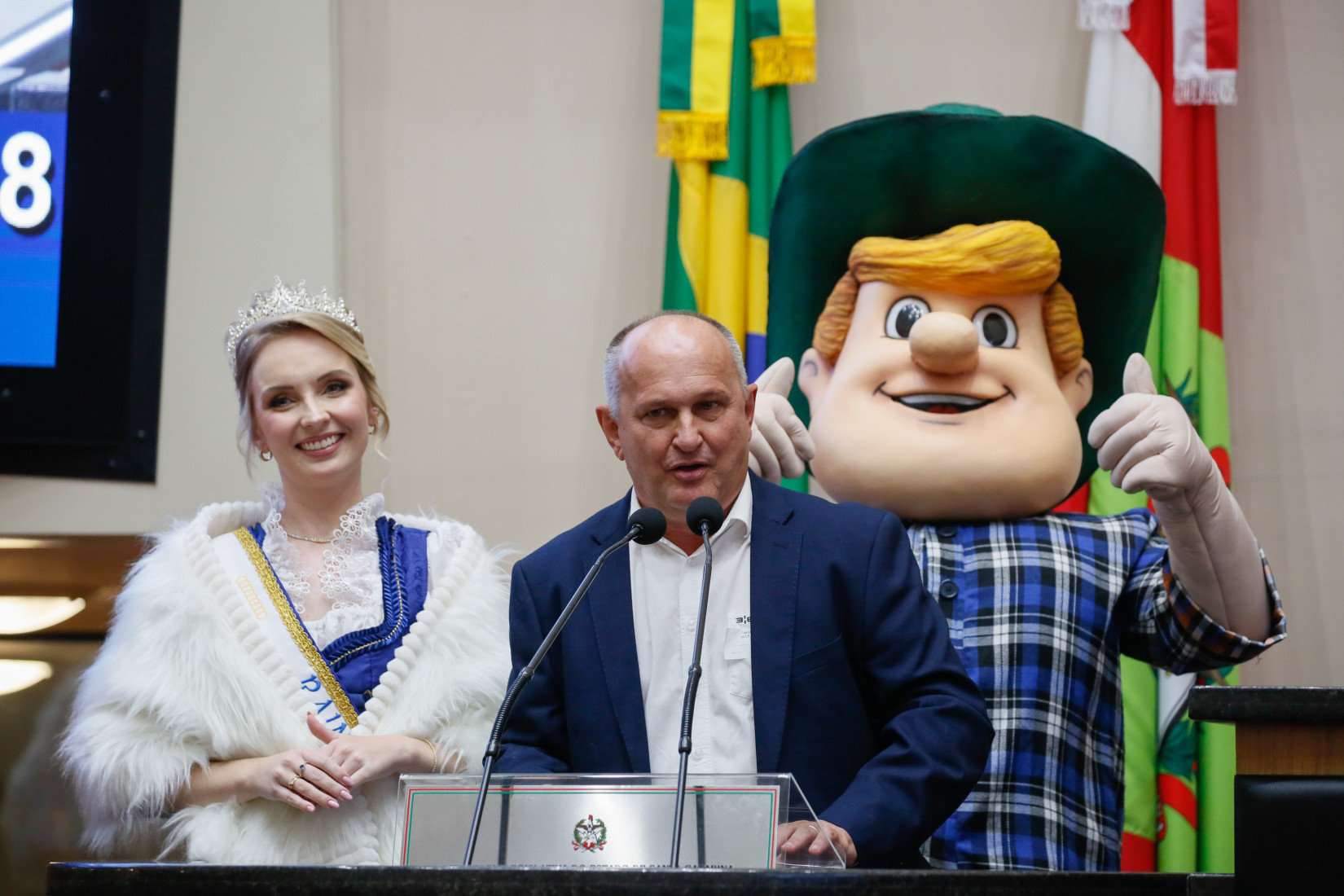 Alceu Gilmar Moretti discursa na tribuna, acompanhado de Anna Letícia Hornburg e do mascote Hans.