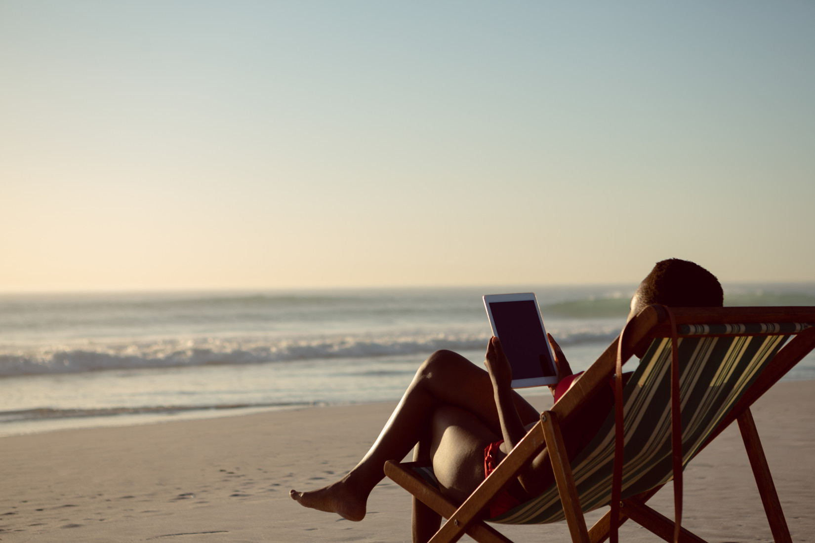 Rear view of woman using digital tablet while relaxing in a beach chair on the beach